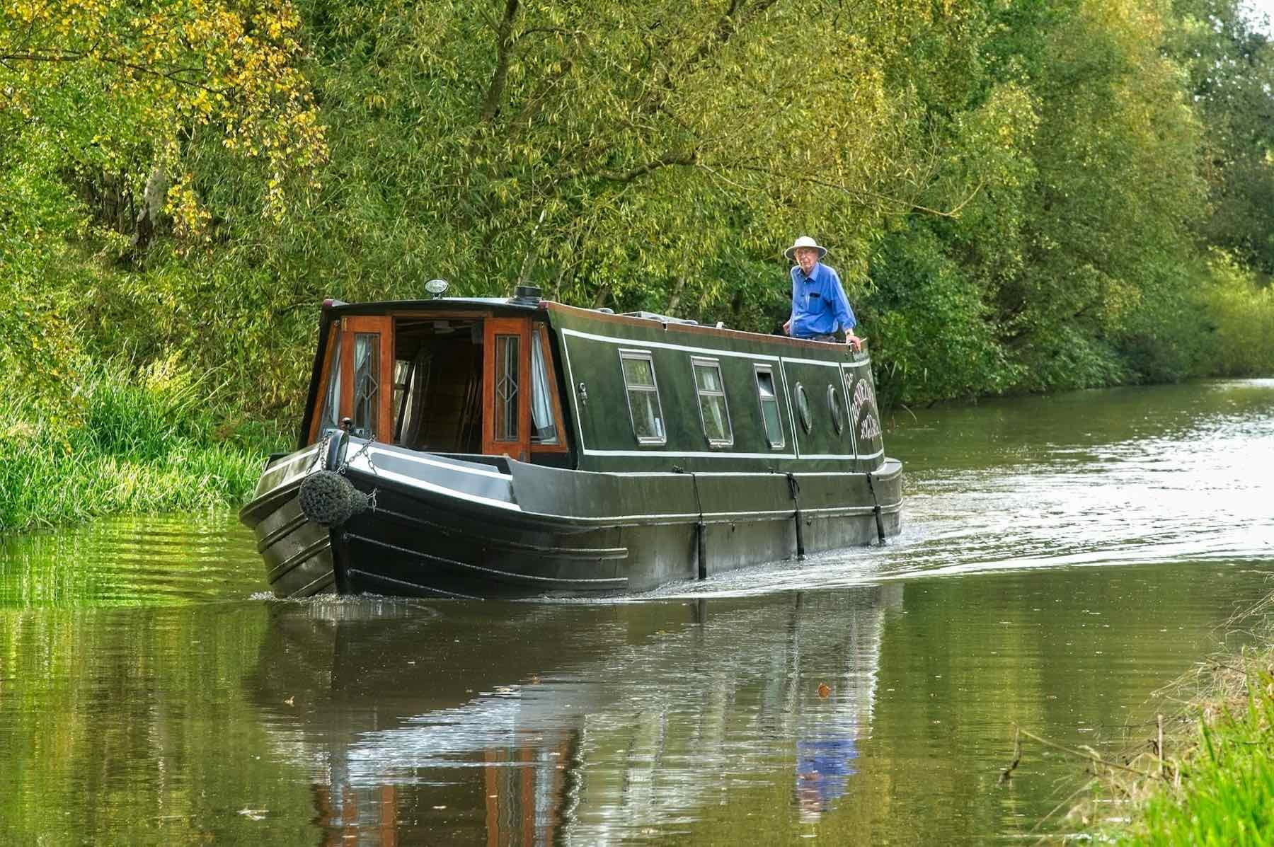 A dark green narrowboat cruising along a tree-lined canal, its steerer standing at the stern
