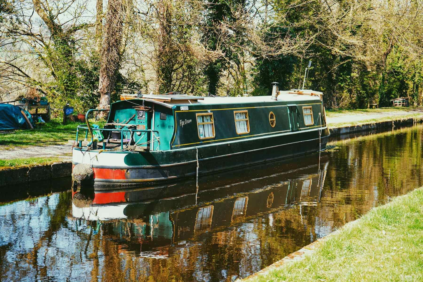 A green narrowboat moored against a grassy towpath, reflected in still water